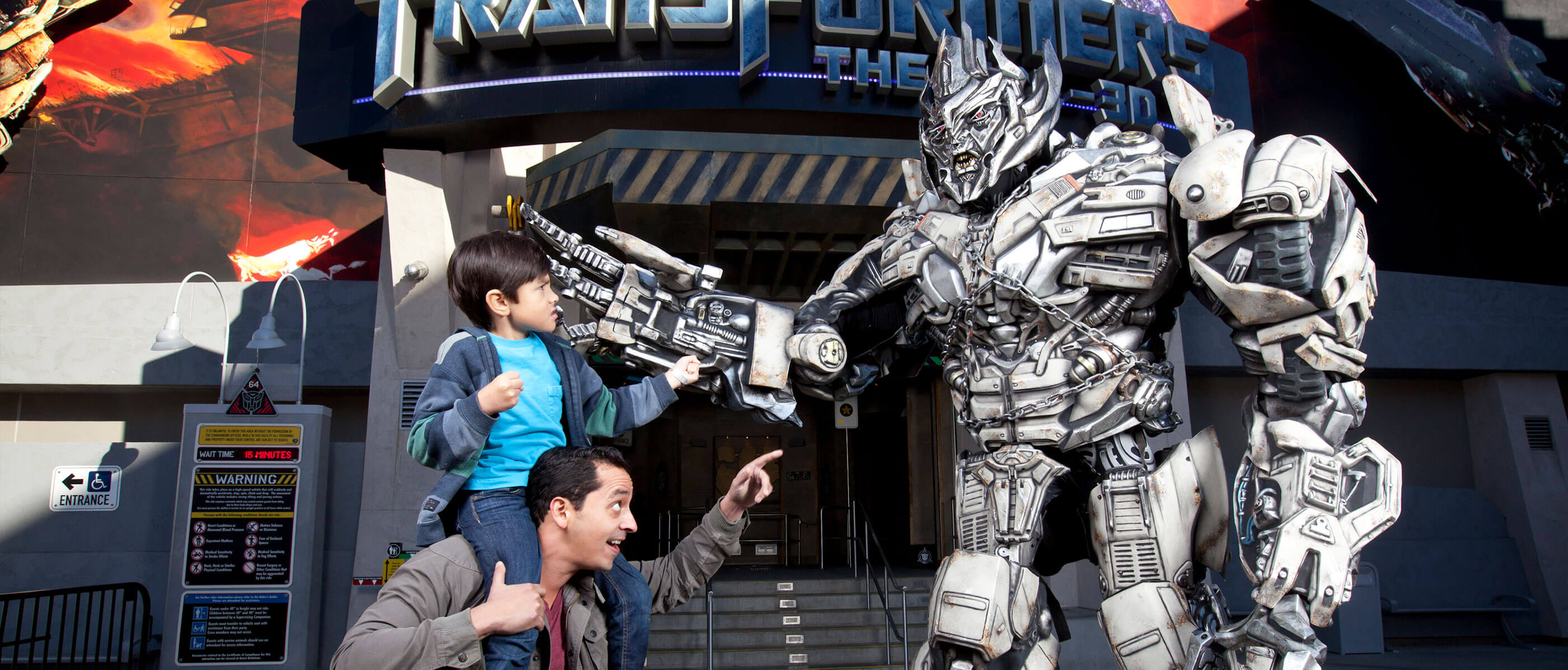 A dad with his son on his shoulders pointing at a transformer in front of the Transformer ride.