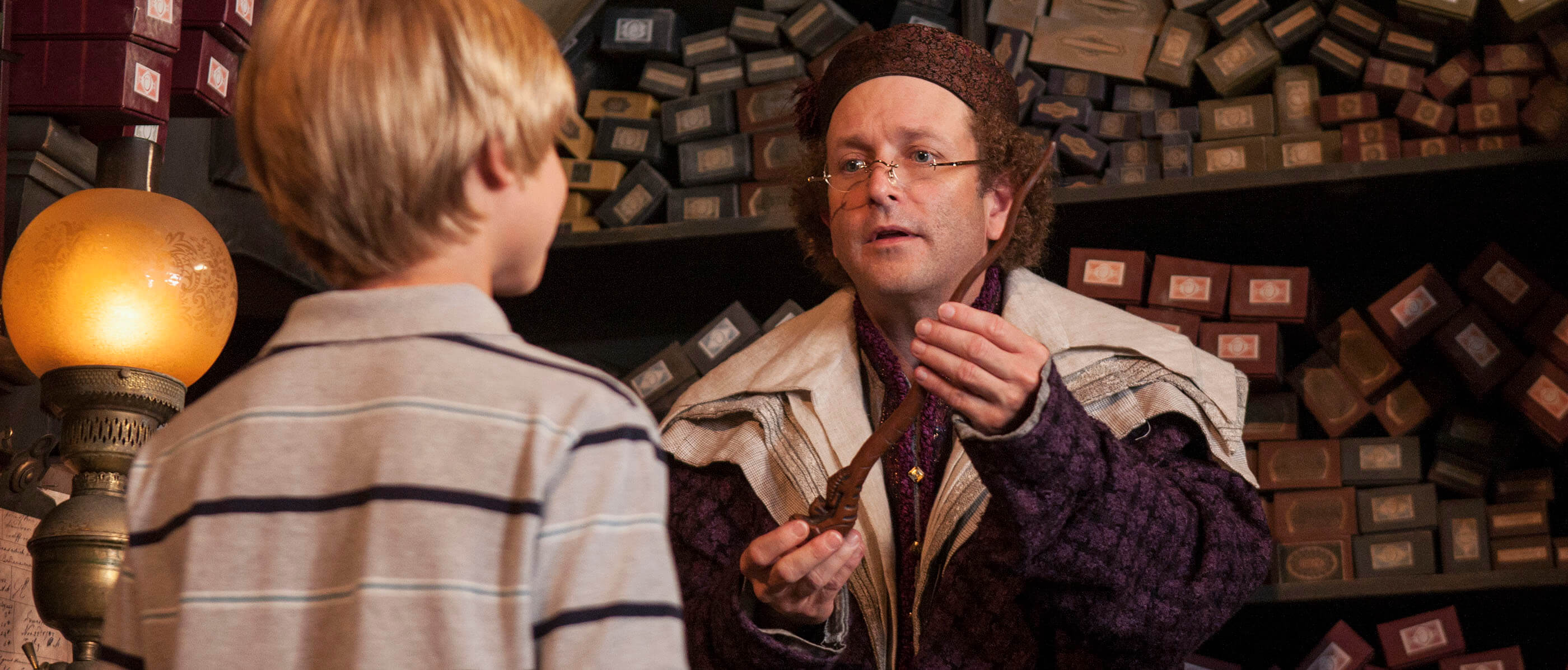 A man dressed up as a wizard in Ollivanders Wand store handing a wand to a young boy.