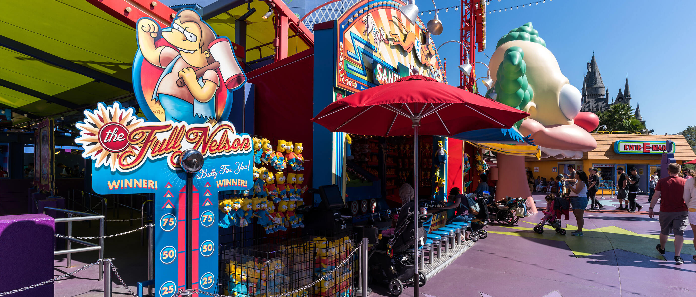 The Fully Nelson game and a red umbrella beside it and people walking about.