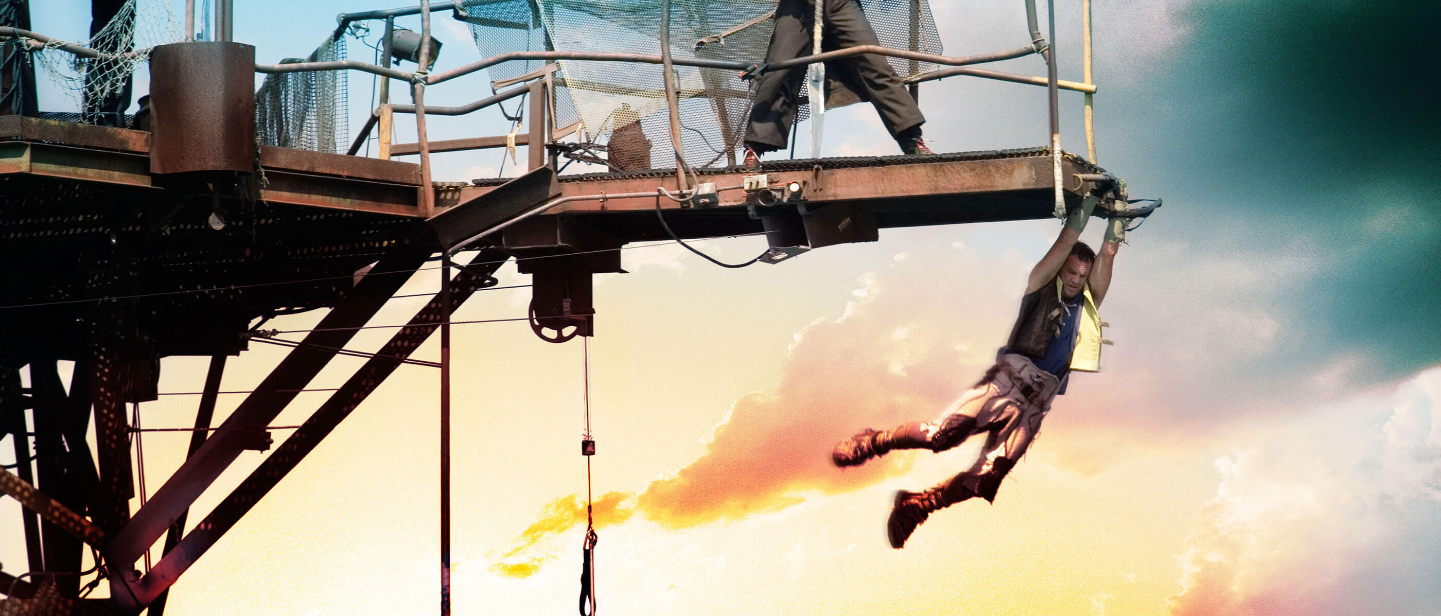 A man hanging from a metal beam edge at sunset.