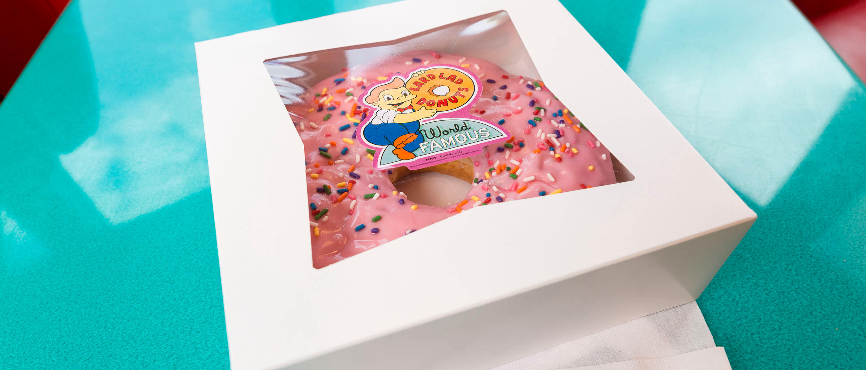 A donut with pink icing, sprinkles and in a Lard Lad Donut box.
