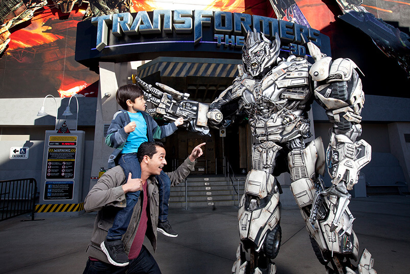 A dad with his son on his shoulders pointing at a transformer in front of the Transformer ride.