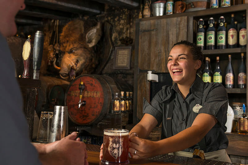 A woman smiling and handing a beer to a customer with a bar behind her.