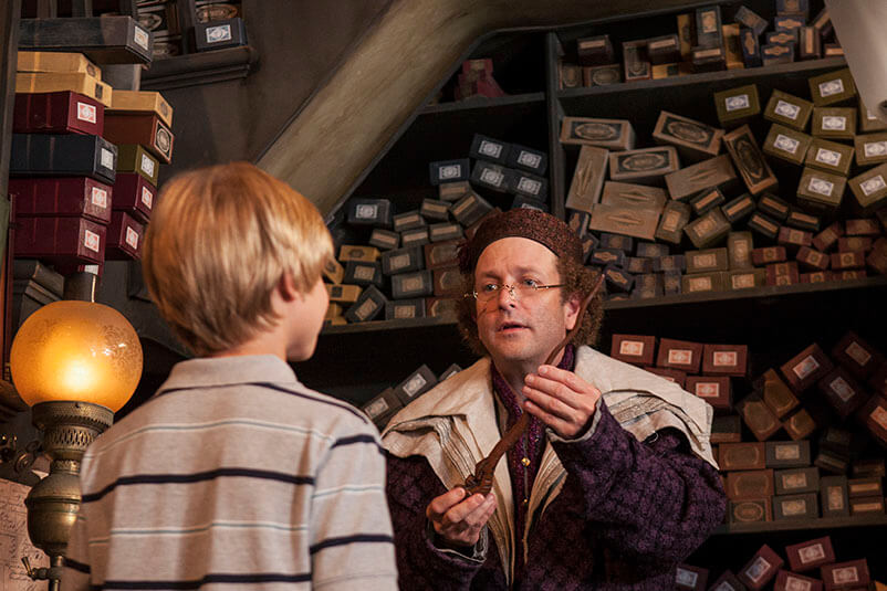 A man dressed up as a wizard in Ollivanders Wand store handing a wand to a young boy.