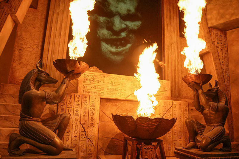 The inside of an Egyptian temple with two figures holding bowls of fire and a green and black figure in the middle.