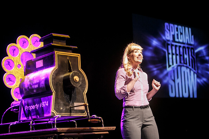 A woman on stage with the Special Effects Show logo in the background and a black object beside her.