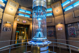 A stone room with a glass figure in a glass case that's place in the middle of the room.