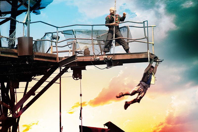 A man hanging from a metal beam edge at sunset.