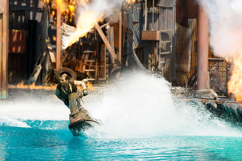 A girl on a jet ski in the water with a building on fire behind.